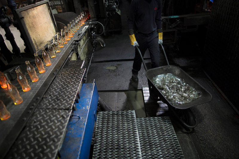 A worker pushes a wheelbarrow with broken glass bottles while new Coke bottles move along the conveyor belt.